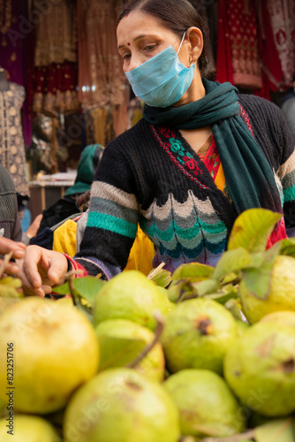 Indian mature woman wearing protective face mask and buying guava from outdoor market 