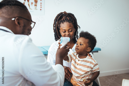 Smiling young African American mother with small 2s boy child son have consultation with caring male pediatrician in private hospital or clinic. Doctor measuring temperature of boy  photo