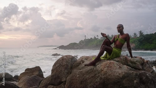 Divine non-binary black person in long open dress brass jewelry poses on rocky hill top above dramatic ocean sunset skyline. Queer lgbtq fashion model in open outfit sits on hill. Pantheon concept photo