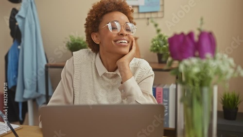 Confident and attractive african american woman joyfully posing and touching hair, smiling fashionably while sitting at home table photo