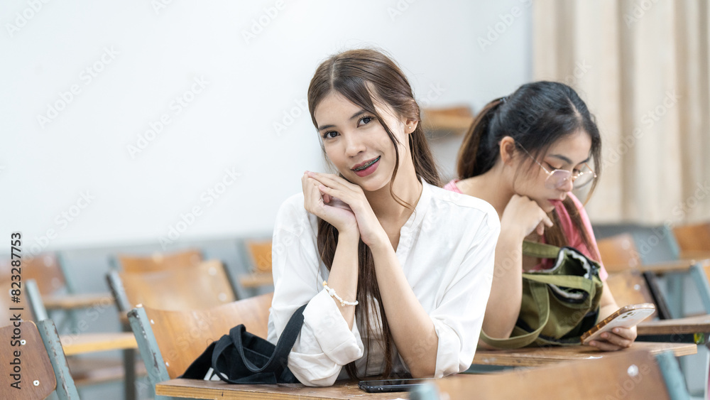 Fototapeta premium Two girls sitting in a classroom, one of them is looking at her phone