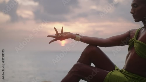 Non-binary person hand touches sun, poses gracefully sitting on top of rock in ocean at sunset in slow motion. Trans ethnic model in open tropical dress on peak of mount wears brass rings with gems. photo