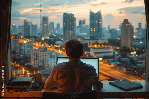 Businessman working late on a laptop in an office with a stunning cityscape view at dusk, showcasing urban lights and skyscrapers.