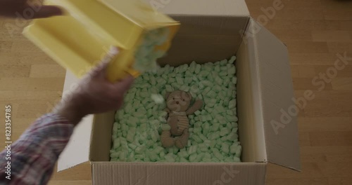 A man in a plaid shirt looks inside a large open cardboard box filled with packing peanuts, discovering a small, fragile item wrapped for protection.