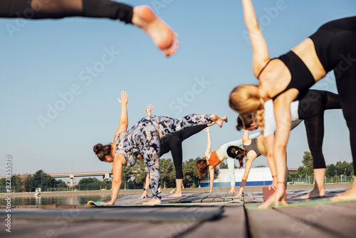 Young women stand in crescent moon pose and do Ardha Chandrasana on pier on river bank. Morning yoga class with teacher. Group of girls is doing physical exercises or Pilates. photo