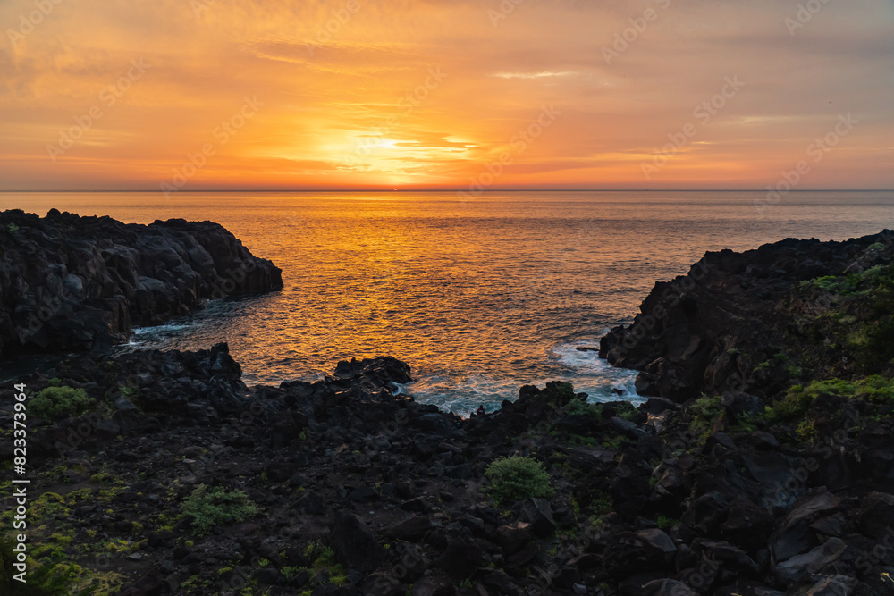 A beautiful coastal area in eastern part of Japan. A clear blue sky and an orange sunrise reflects on the sea water. A big rock formation in the coast line. Island from the distance. A beautiful coastal area in eastern part of Japan. A clear blue sky and an orange sunrise reflects on the sea water. A big rock formation in the coast line. Island from the distance.