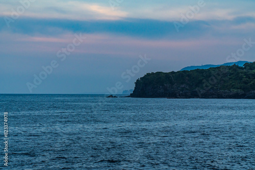A beautiful coastal area in eastern part of Japan. A clear blue sky and an orange sunrise reflects on the sea water. A big rock formation in the coast line. Island from the distance.
