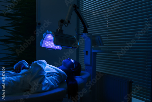 Remote view of female patient undergoes blue LED light therapy for skin rejuvenation at modern aesthetic clinic. Using phototherapy device with blue light wavelength for acne treatments.
