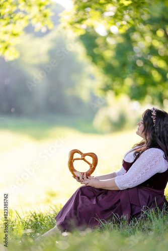 beautiful, young, bavarian woman in traditional costume, dirndl, sitting in nature on the meadow in front of the oktoberfest with a pretzel 