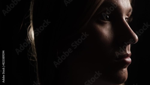 Thoughtful, determined face of woman against black background in spotlight. Camera pans around woman with purposeful gaze as beautiful spotlights appear in background. Close-up photo