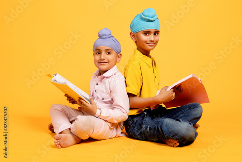 Portrait of Two smiling indian sikh school students sitting on floor holding note books studying isolated over yellow background. students preparing for exams, Knowledge and intelligence, education. photo