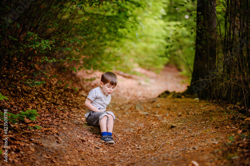 A young boy sits on a leaf-covered forest path, looking back towards the camera. Surrounded by lush green foliage, he appears curious and contemplative. © Иванна Емельянова