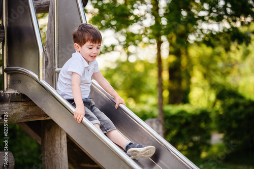 A happy young boy slides down a metal playground slide on a sunny day, surrounded by lush green trees, enjoying outdoor playtime.