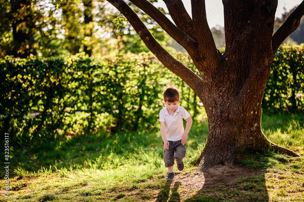 A young boy in a white shirt and denim shorts runs near a large tree in a park, enjoying the lush greenery and sunlight on a summer day.