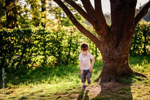 A young boy in a white shirt and denim shorts runs near a large tree in a park, enjoying the lush greenery and sunlight on a summer day. © Иванна Емельянова