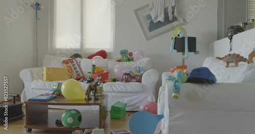 A living room shows the aftermath of a children's party, with toys and balloons spread across white couches and the floor.