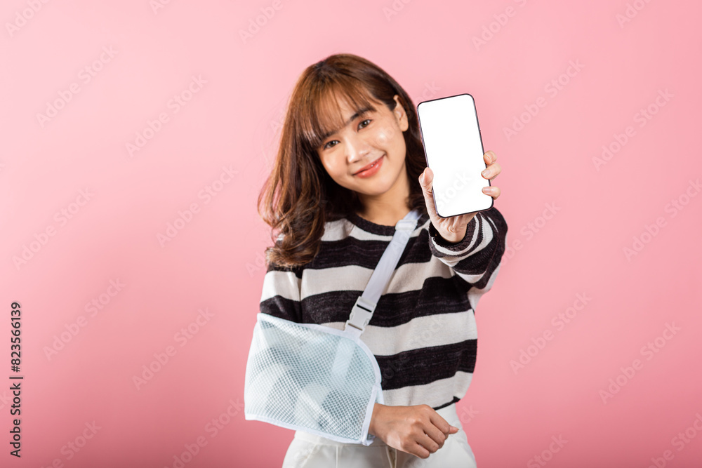 Naklejka premium Happy Asian woman with a broken arm, wearing an arm splint, smiles while displaying her smartphone screen. Coping with pain and recovery, isolated on pink.