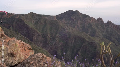 Pan shot of two paragliders flying by in beautiful landscape mountains of tenerife island national park near City of Adeje. Filmed from a mountain top following the persons as the move by to the ocean