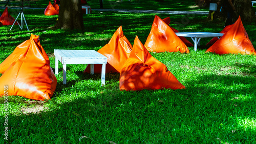 Beautiful view of orange triangle bean bags and cushions with white wooden table for customers to relax outdoors on green grass under shade of large tree with blurred background. Selective focus.