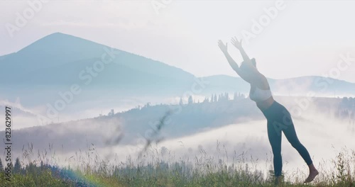 Slim woman practicing daylight yoga at mountains in early morning. Side view of graceful female wearing activewear, doing Dancer pose of yoga, in morning sunlight outdoors. Concept of yoga. photo