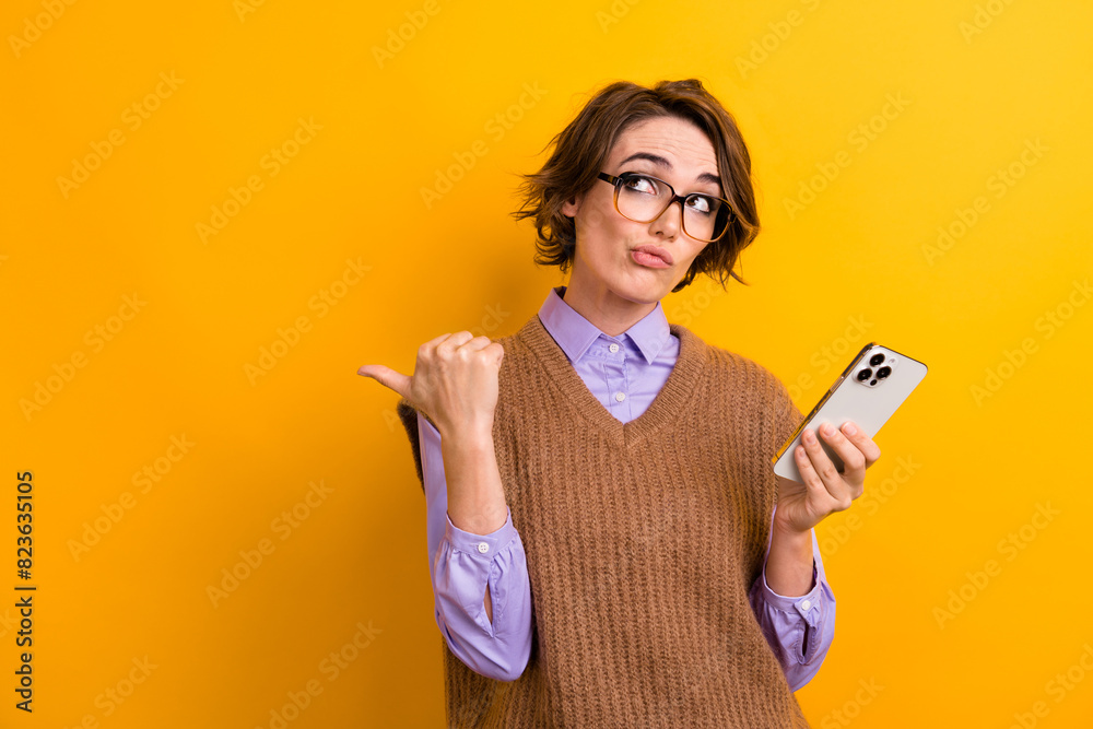 Portrait of attractive woman wear knit waistcoat hold smartphone look directing at offer empty space isolated on yellow color background Portrait of attractive woman wear knit waistcoat hold smartphone look directing at offer empty space isolated on yellow color background