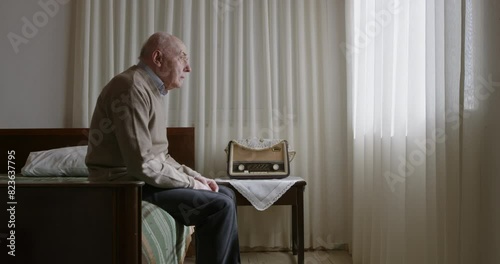 A senior man sits on his bed, beside a large window, gazing thoughtfully at a vintage radio in a peaceful room.