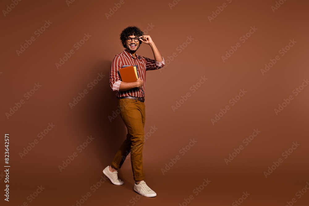 Full body portrait of nice young man hold book walk empty space wear striped shirt isolated on brown color background Full body portrait of nice young man hold book walk empty space wear striped shirt isolated on brown color background