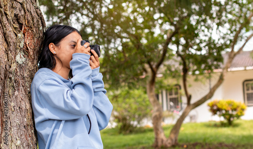 Photography lifestyle: young woman leaning against a tree taking pictures with analog camera