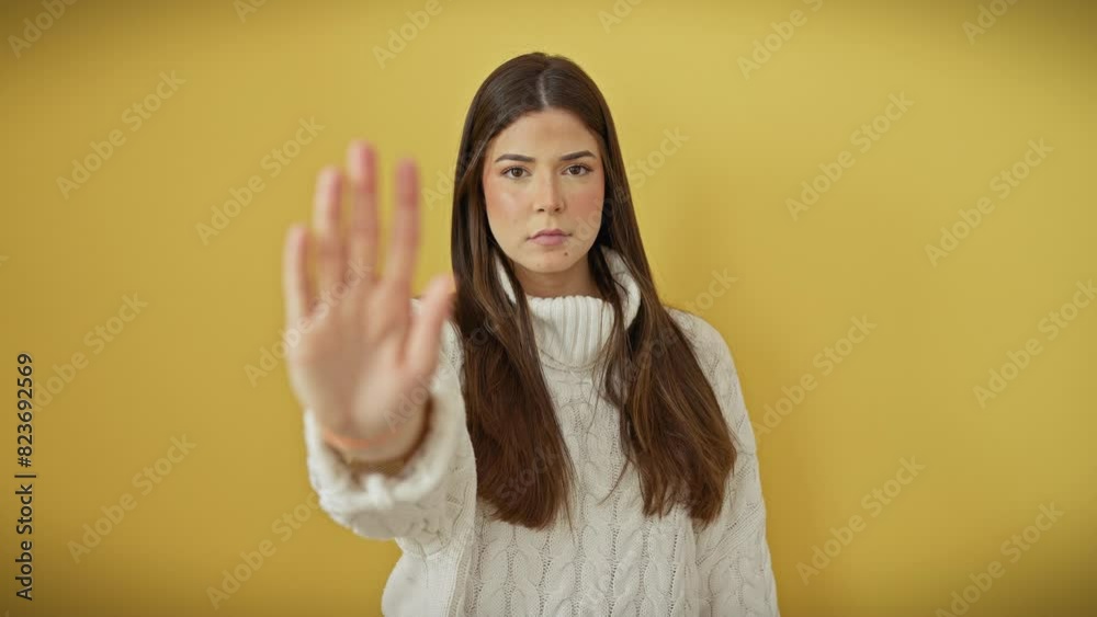 Serious young hispanic woman in sweater, confidently projecting a stop gesture smartly with palm, expressing negative no-go warning over yellow isolated cutout Serious young hispanic woman in sweater, confidently projecting a stop gesture smartly with palm, expressing negative no-go warning over yellow isolated cutout