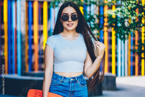 Portrait of attractive brunette young woman with gorgeous brunette hair smiling at camera while holding skateboard in hands for spending summer free time on hobby.Teen in sunglasses standing outdoors © BullRun
