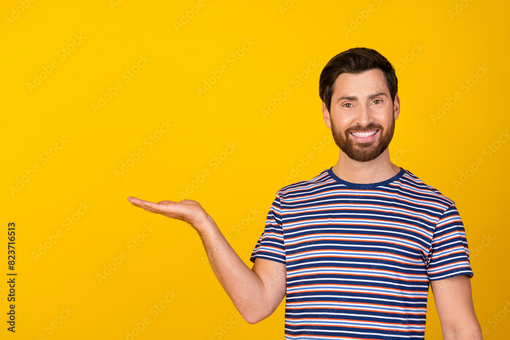 Photo of ecstatic man with stylish beard dressed striped t-shirt palm presenting product empty space isolated on yellow color background Photo of ecstatic man with stylish beard dressed striped t-shirt palm presenting product empty space isolated on yellow color background