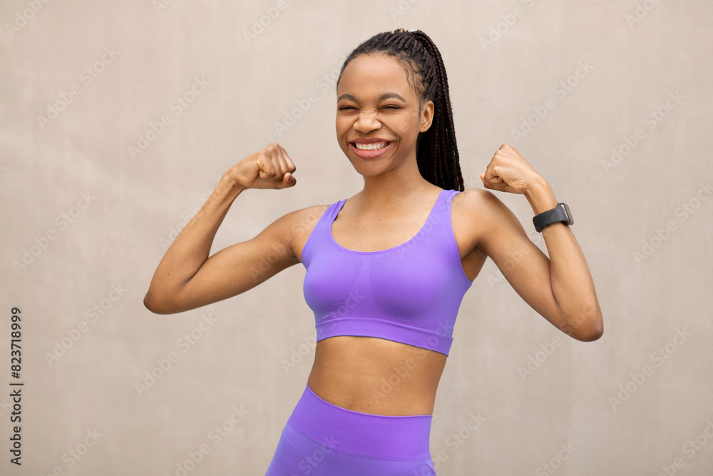 Young slim black woman in sportswear showing her biceps and laughing, posing outdoors over beige wall after workout, showcasing healthy lifestyle Young slim black woman in sportswear showing her biceps and laughing, posing outdoors over beige wall after workout, showcasing healthy lifestyle