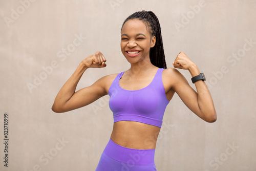 Young slim black woman in sportswear showing her biceps and laughing, posing outdoors over beige wall after workout, showcasing healthy lifestyle