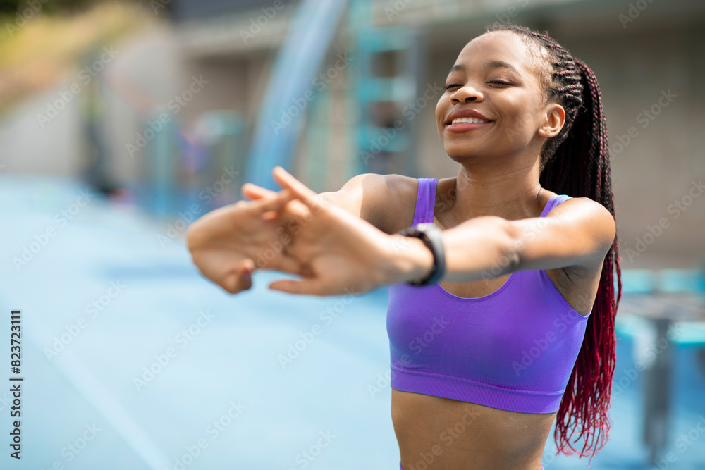 Smiling black lady stretching arms before training outdoors, warming up her hands during outside workout on sports ground, copy space Smiling black lady stretching arms before training outdoors, warming up her hands during outside workout on sports ground, copy space