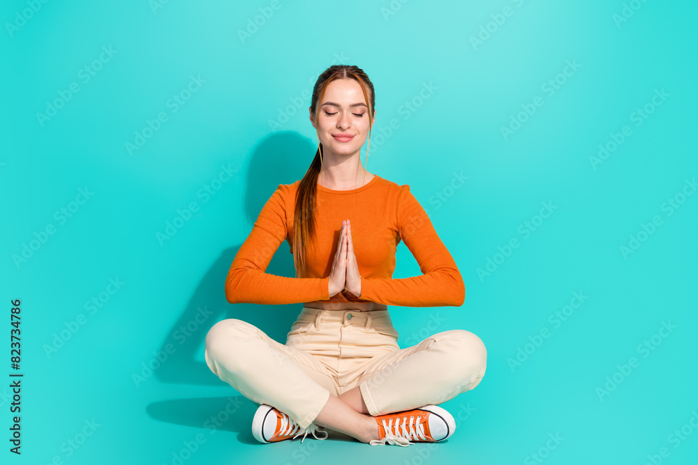 Full size photo of appreciative girl dressed orange shirt white trousers sit on floor palms together isolated on teal color background Full size photo of appreciative girl dressed orange shirt white trousers sit on floor palms together isolated on teal color background