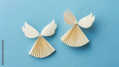 two paper angel ornaments isolated on a blue background  with a simple style and design using minimalistic shapes and simple colors of beige and white in a flat lay.