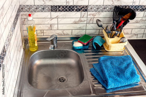 Kitchen sink with utensils in yellow holder, dish soap and sponges on side. Stainless steel basin next to blue dish towel, tiled backdrop