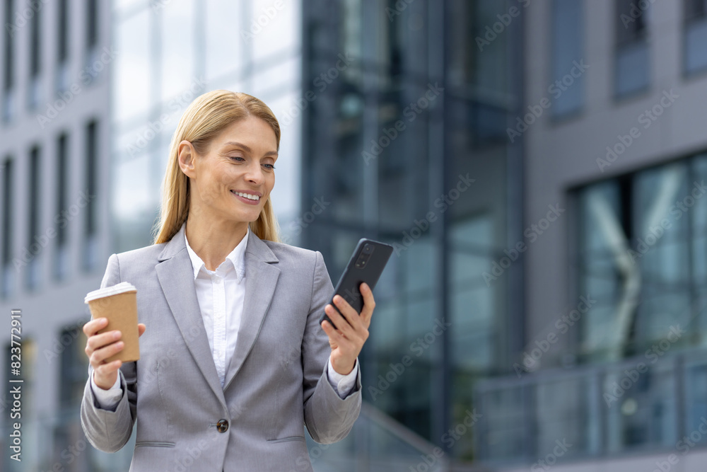 Fototapeta premium In a modern city, a professional businesswoman in a grey suit smiles while checking her phone and holding a coffee cup outside an office building. She exudes confidence and success
