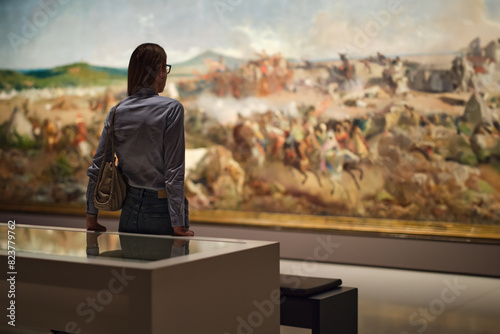 Thoughtful young Caucasian woman wearing glasses and looking at exhibition. Concept of Museum Day.