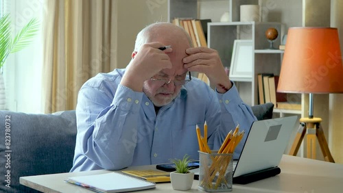 An elderly businessman, gray-haired in a blue shirt, at his desk, working on a computer, reconciling the data of an accounting report. Tired and falling asleep. Slow motion. photo