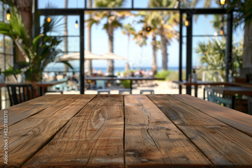 A wooden table in the foreground with a blurred background of a beachside cafe. The background shows sandy beach views through large windows  casual seating  tropical plants  and customers