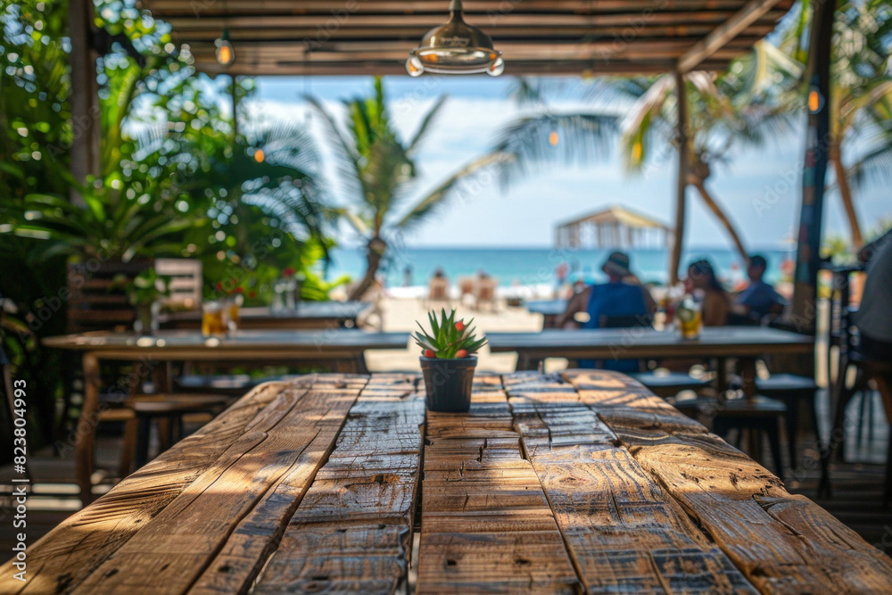 A wooden table in the foreground with a blurred background of a beachside cafe. The background shows sandy beach views through large windows, casual seating, tropical plants, and customers A wooden table in the foreground with a blurred background of a beachside cafe. The background shows sandy beach views through large windows, casual seating, tropical plants, and customers
