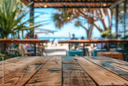 A wooden table in the foreground with a blurred background of a beachside cafe. The background shows sandy beach views through large windows  casual seating  tropical plants  and customers