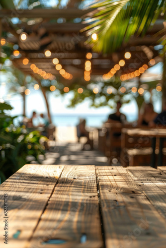A wooden table in the foreground with a blurred background of a beachside cafe. The background shows sandy beach views through large windows, casual seating, tropical plants, and customers © grey