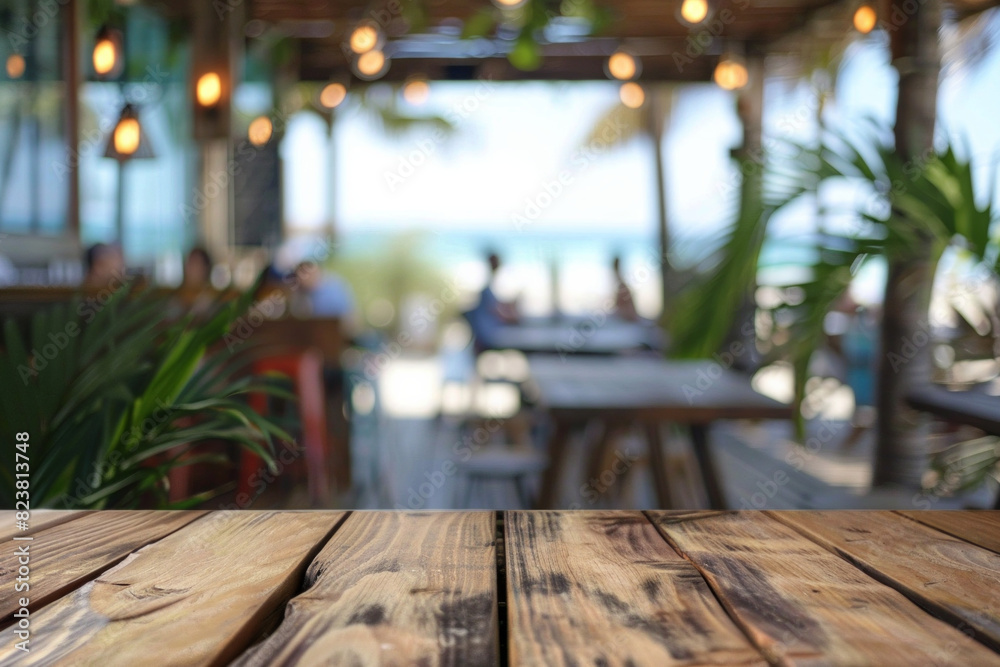 A wooden table in the foreground with a blurred background of a beachside cafe. The background shows sandy beach views through large windows, casual seating, tropical plants, and customers A wooden table in the foreground with a blurred background of a beachside cafe. The background shows sandy beach views through large windows, casual seating, tropical plants, and customers