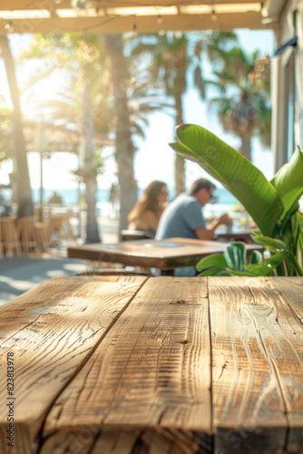 A wooden table in the foreground with a blurred background of a beachside cafe. The background shows sandy beach views through large windows, casual seating, tropical plants, and customers