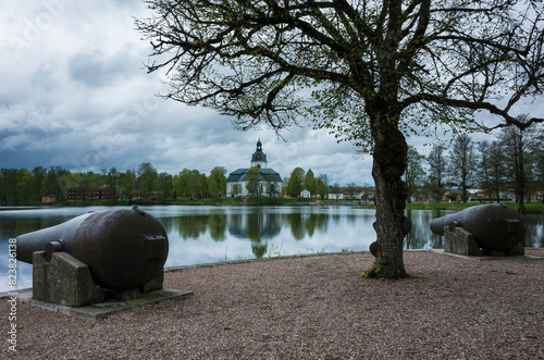 Filipstad's Church and cannons on Kanonudden in Filipstad, White church building in the distance is reflecting in the water, Two large cannons, A dark tree with small leaves, Cloudy weather photo
