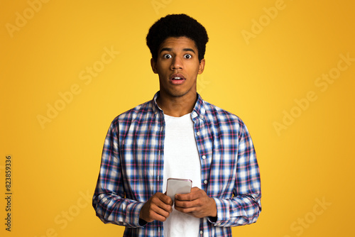 Shocked african-american guy texting on phone, looking at camera over orange background