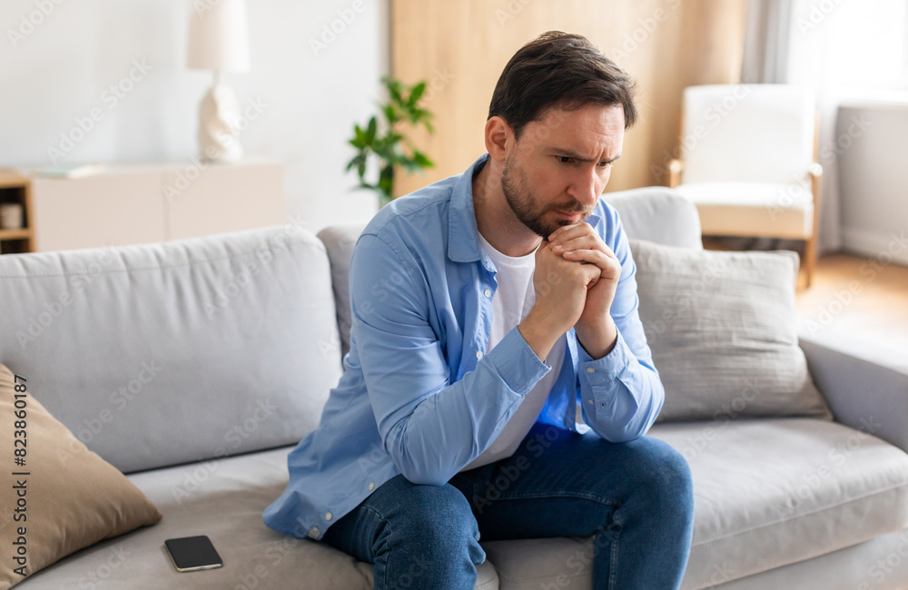 A man is seated on a couch, deep in thought, with his hand resting on his chin. He appears contemplative and engaged in introspection. A man is seated on a couch, deep in thought, with his hand resting on his chin. He appears contemplative and engaged in introspection.