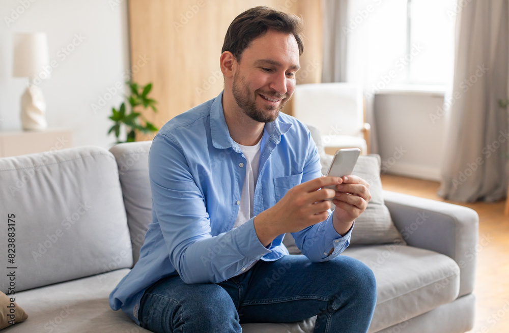 A bearded man is seated on a couch, engrossed in his smartphone screen. He appears focused as he scrolls and interacts with the device, surrounded by a living room setting. A bearded man is seated on a couch, engrossed in his smartphone screen. He appears focused as he scrolls and interacts with the device, surrounded by a living room setting.